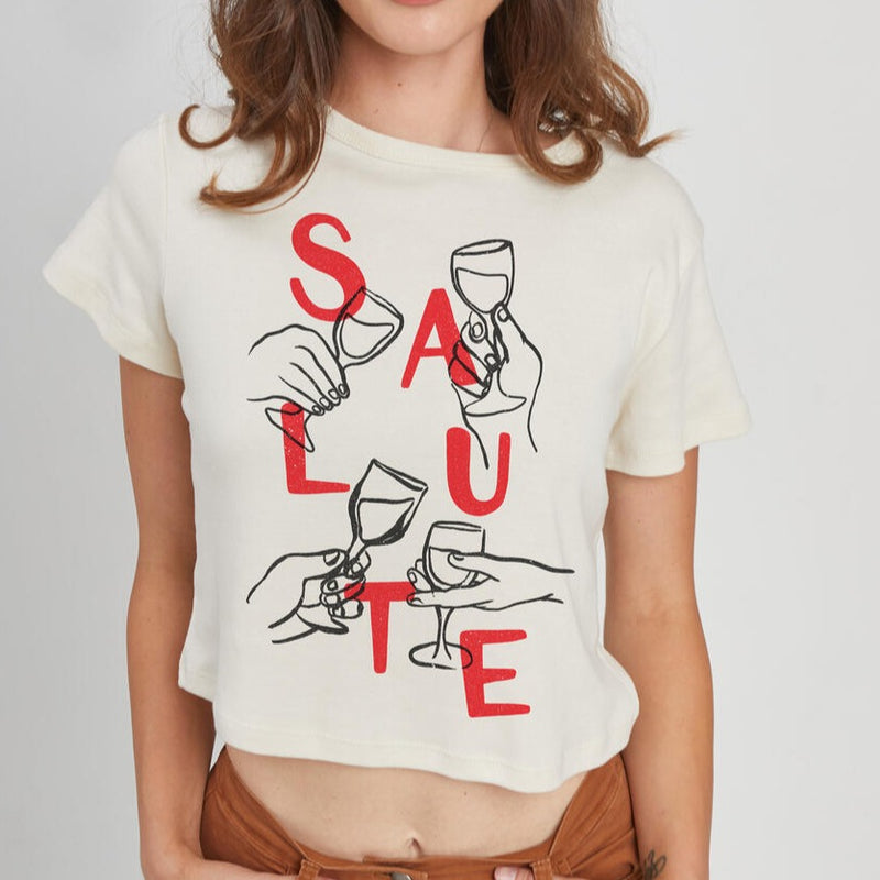 front view of model wearing the salute baby tee. shows the crew neckline. also shows the short sleeves, the cropped length and the front graphic with hands cheering with glasses and it says salute. 