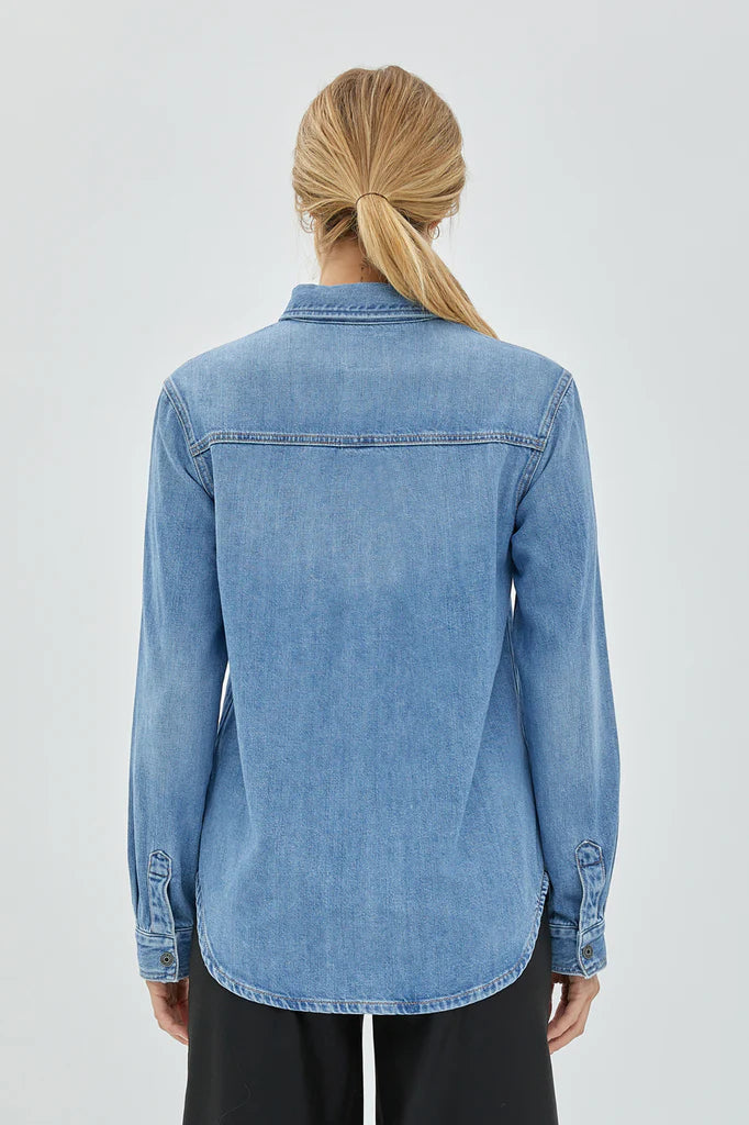 back view of the model wearing the abbey denim shirt. shows the button closures on the sleeves. also shows the collared neckline, the rounded bottom hem, and the seaming across  the top shoulder. 