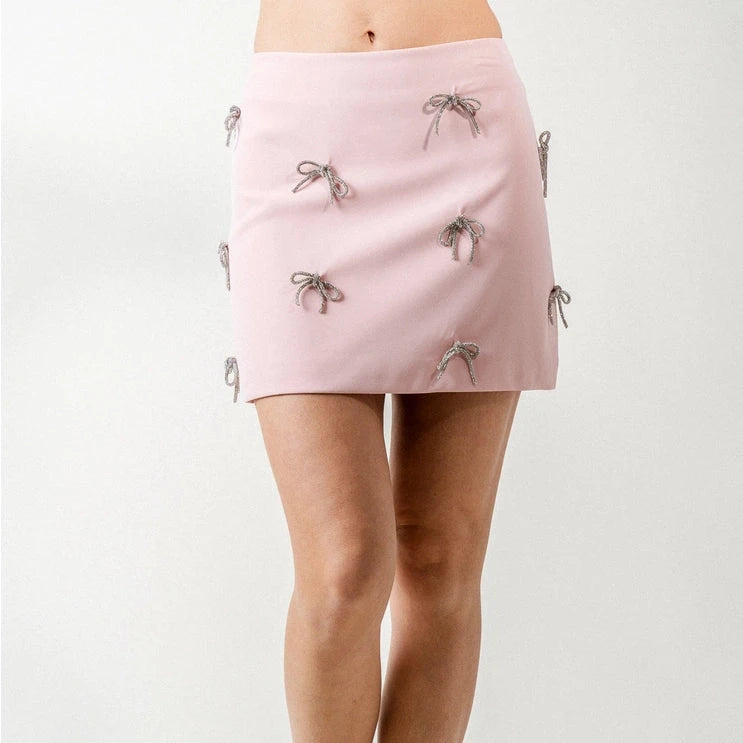 front view of the model wearing the sabrina rhinestone bow mini skirt. shows the mid-rise. also shows the mini length, the light pink color and the rhinestone bow details. 