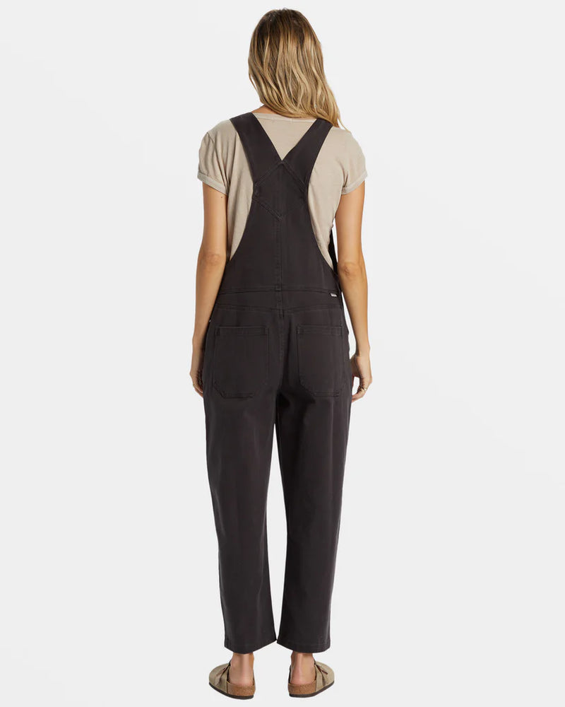 back view of the model wearing the sand canyon overalls. shows the black sands color. also shows the back patch pockets, the relaxed fit and the straps. 