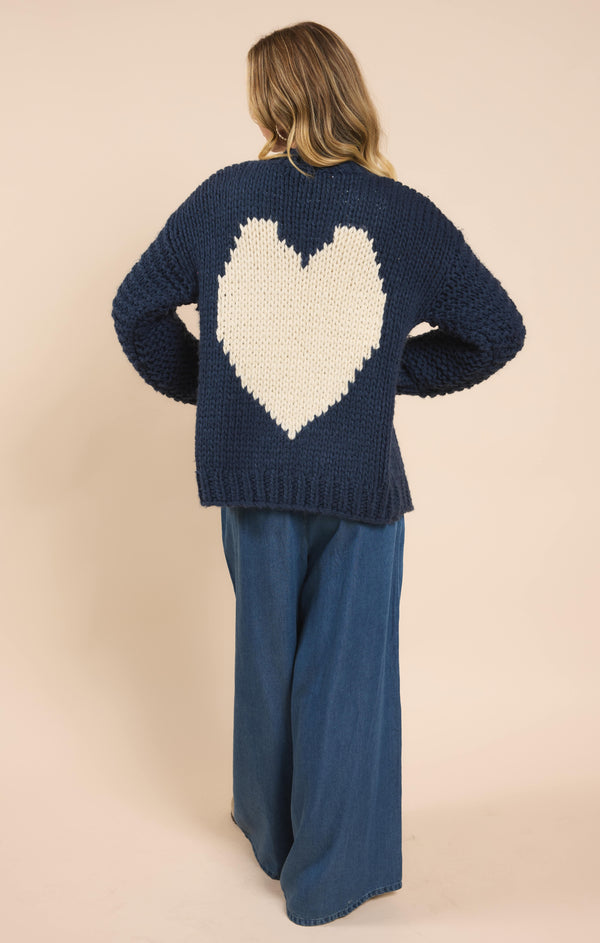 back view of the model wearing the heart you handknit cardigan. shows the ribbed detailing. also shows the ivory heart detail, the mid length and the navy color of the cardigan. 