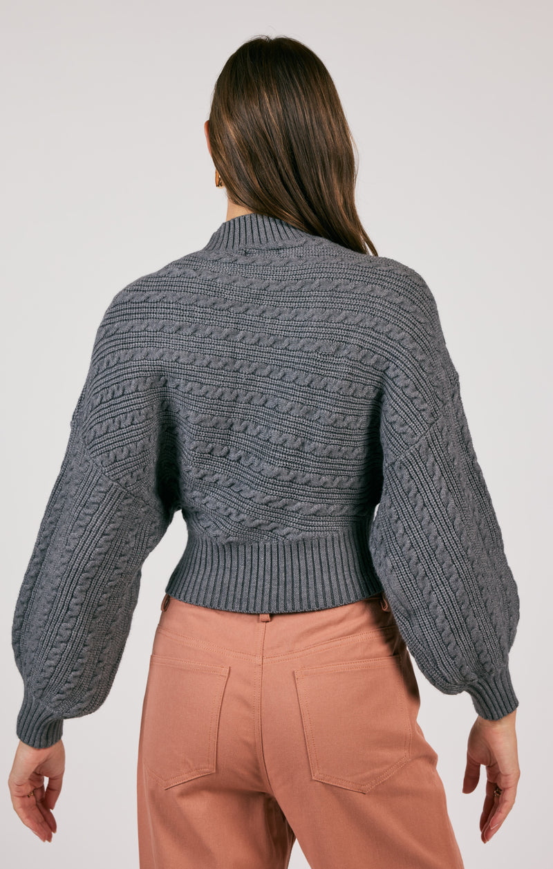 back view of the model wearing the along the vines cable knit sweater. shows the cropped length. also shows the ribbed detailing, the cable knit detailing throughout, and the exaggerated sleeves.  