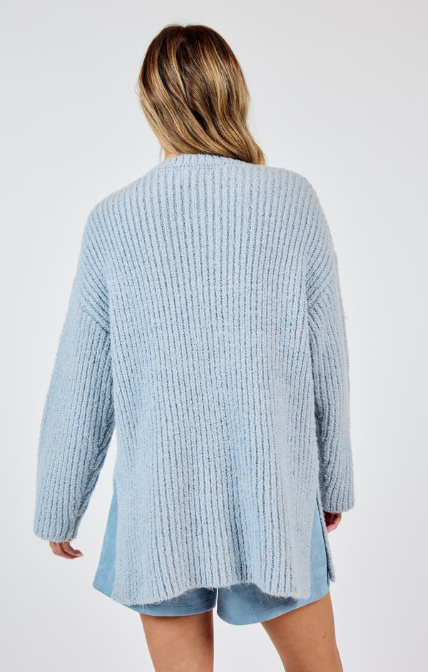 back view of the model wearing the your needs sweater. shows the ribbed detailing. also shows the oversized fit, dropped shoulders and side slits. 