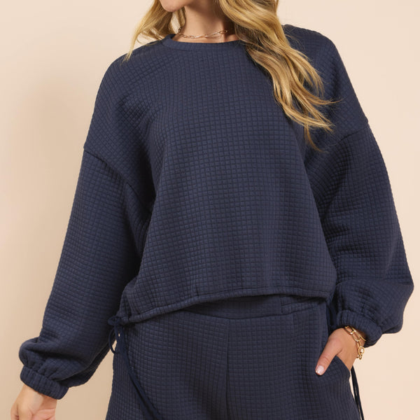 front view of the model wearing the pastime textured pullover. shows the crew neckline. also shows the dropped shoulders, the side ties, the textured pattern. its in this navy color. 