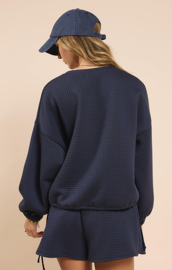 back view of the model wearing the pastime textured pullover. shows the dropped shoulders. also shows the cinched waistband, and the textured detailing. its in this navy color. 